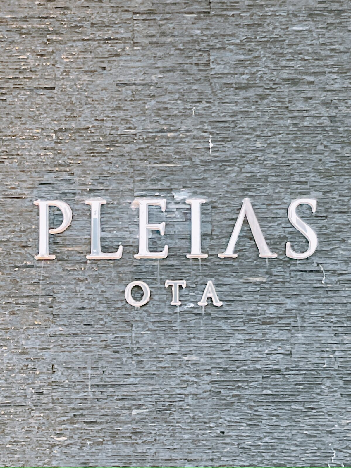 石を積んだ壁に、PLEIAS OTA と白い文字で書かれた看板