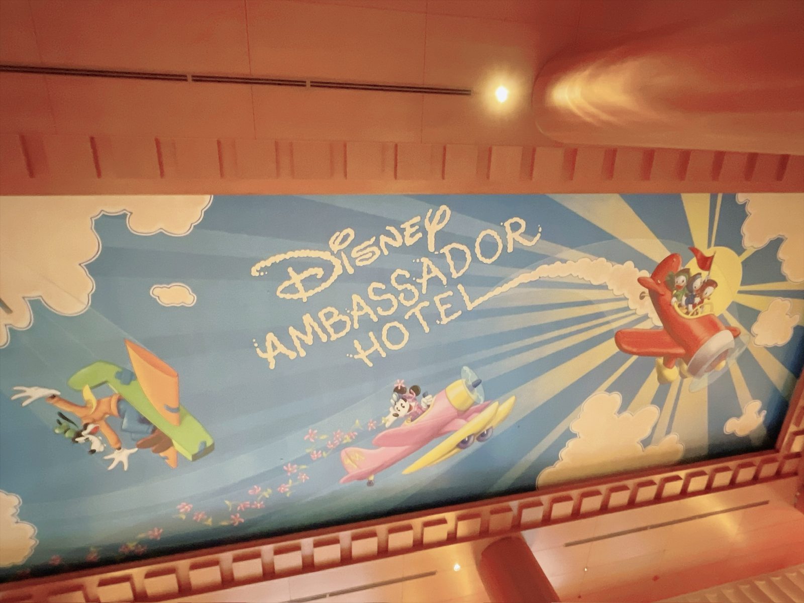 ロビーの天井に描かれた青空の絵。雲の文字で「Disney AMBASSADOR HOTEL」と書かれ、緑の飛行機に乗ったグーフィー、ピンクの飛行機に乗ったミニー、太陽を背にした赤い飛行機に乗った3羽のアヒルの子が飛んでいる。まわりは白い雲とピンクの小花
