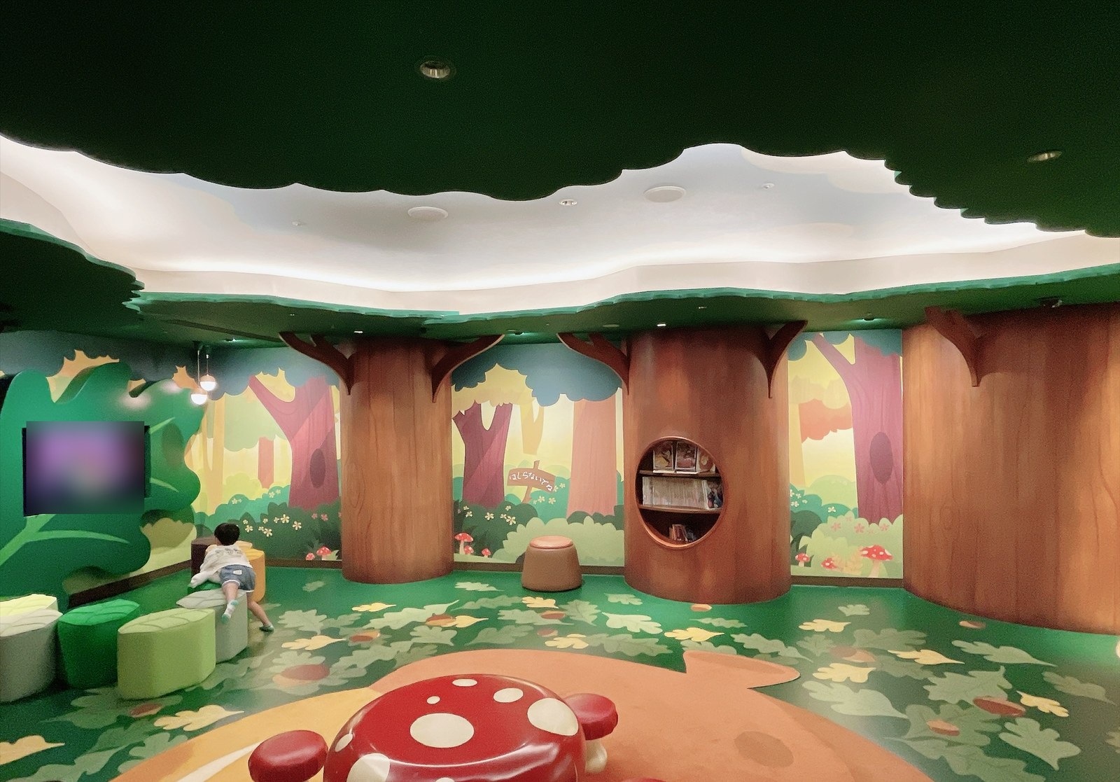 森をテーマにした部屋の全体。木の葉の形の天井と、森の壁画。中央にきのこの形のテーブルと椅子、木の幹には絵本棚がある。左の壁の大きなテレビは画面をぼかし処理。手前に後ろ向きの子どもが写っている(顔は写っていません)