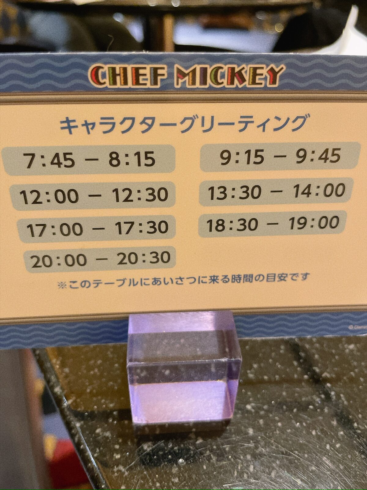 テーブルに立てられた小さな案内。上に「CHEF MICKEY」、その下に「キャラクターグリーティング」と書かれ、7:45-8:15／9:15-9:45／12:00-12:30／13:30-14:00／17:00-17:30／18:30-19:00／20:00-20:30の時間が並ぶ。いちばん下に「このテーブルにあいさつに来る時間の目安です」とある