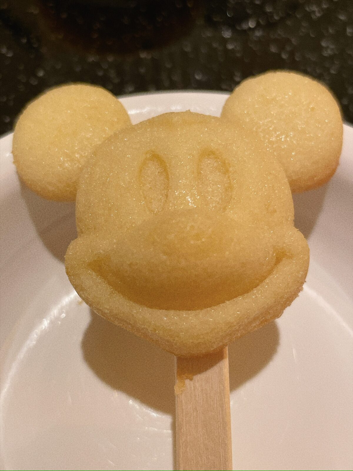 白い皿の上の、ミッキーの顔の形に焼いた薄黄色の生地。目・鼻・口の線が浮き出ていて、下に木の棒が刺さっている