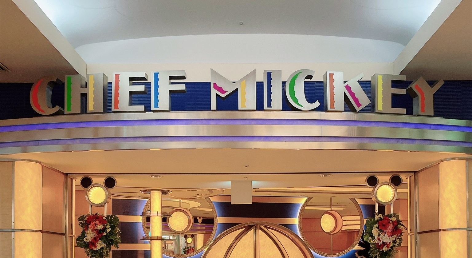 紺色の壁に「CHEF MICKEY」の文字が、赤・緑・黄・青・ピンクなど1文字ずつ違う色で並ぶ看板。下に丸いミッキーの形をしたライトが左右に2つずつ並び、入口の両脇に赤と白の花が飾られている