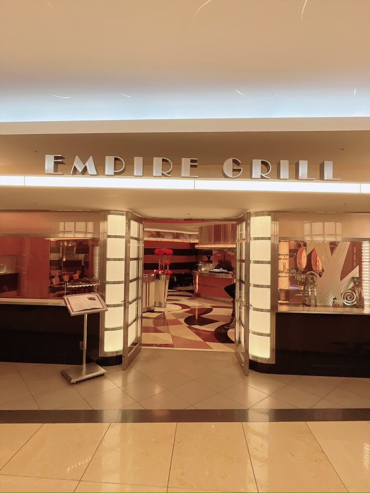 銀色のアールデコ調の看板に「EMPIRE GRILL」の文字。下にガラスの両開きドアが開いていて、左右に光る柱状のパネルが並ぶ。手前左にメニューを載せたスタンドがあり、床はベージュとえんじ色の渦模様のタイル。奥に赤い壁と、赤い花を生けた花瓶が見える