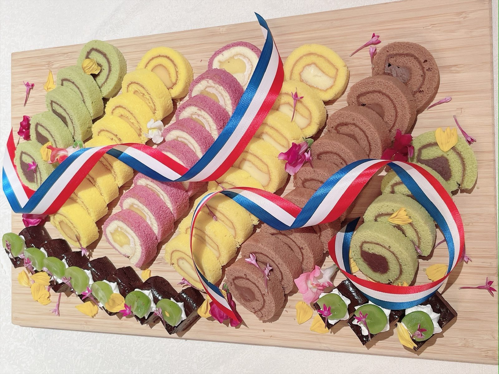 木の板の上に、抹茶・黄色・ピンク・チョコレートなど色とりどりのロールケーキが並び、三色のリボンと花で飾られている