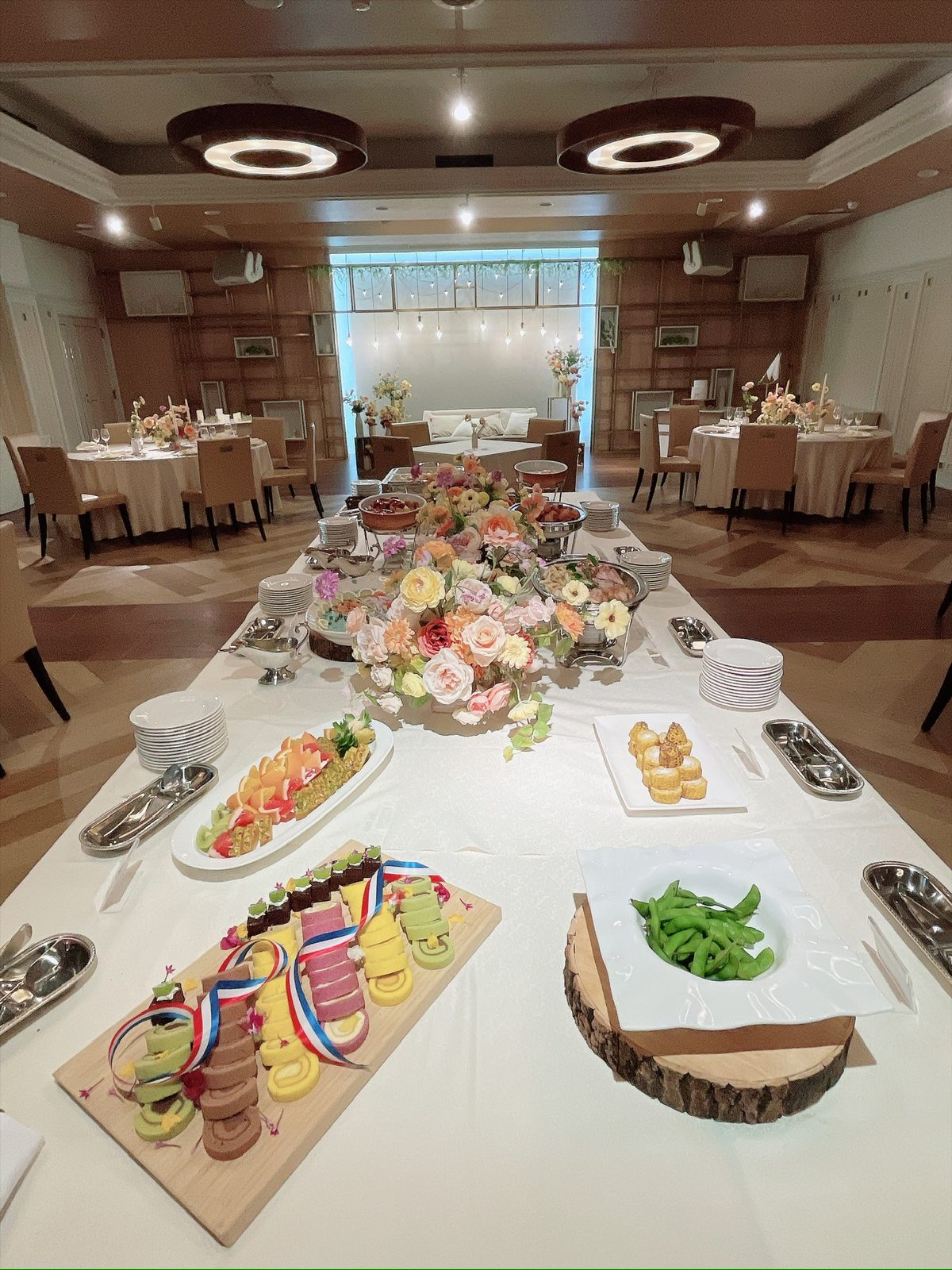 会場中央の長いテーブルに花で飾られた料理が並び、奥に白いクロスの円卓とソファ席が見える