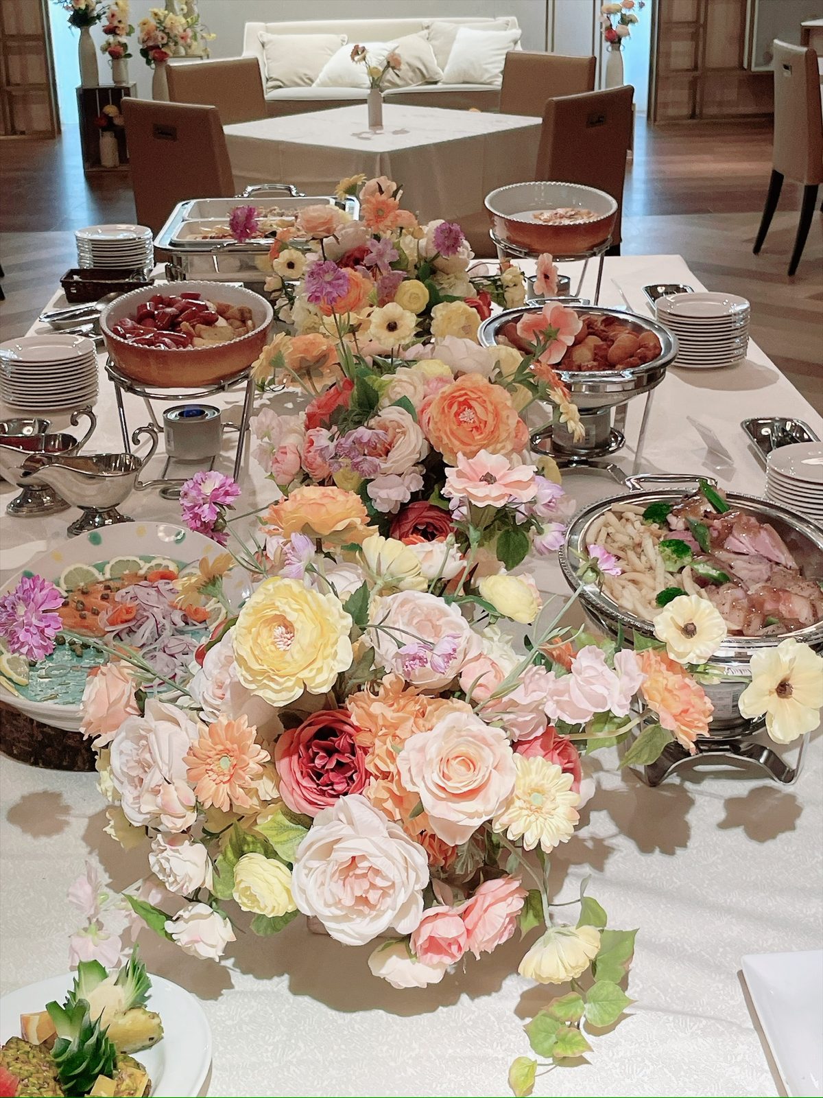 長いテーブルの中央にオレンジやピンクの花が連なる装飾。周りに料理の皿や銀の器が並んでいる