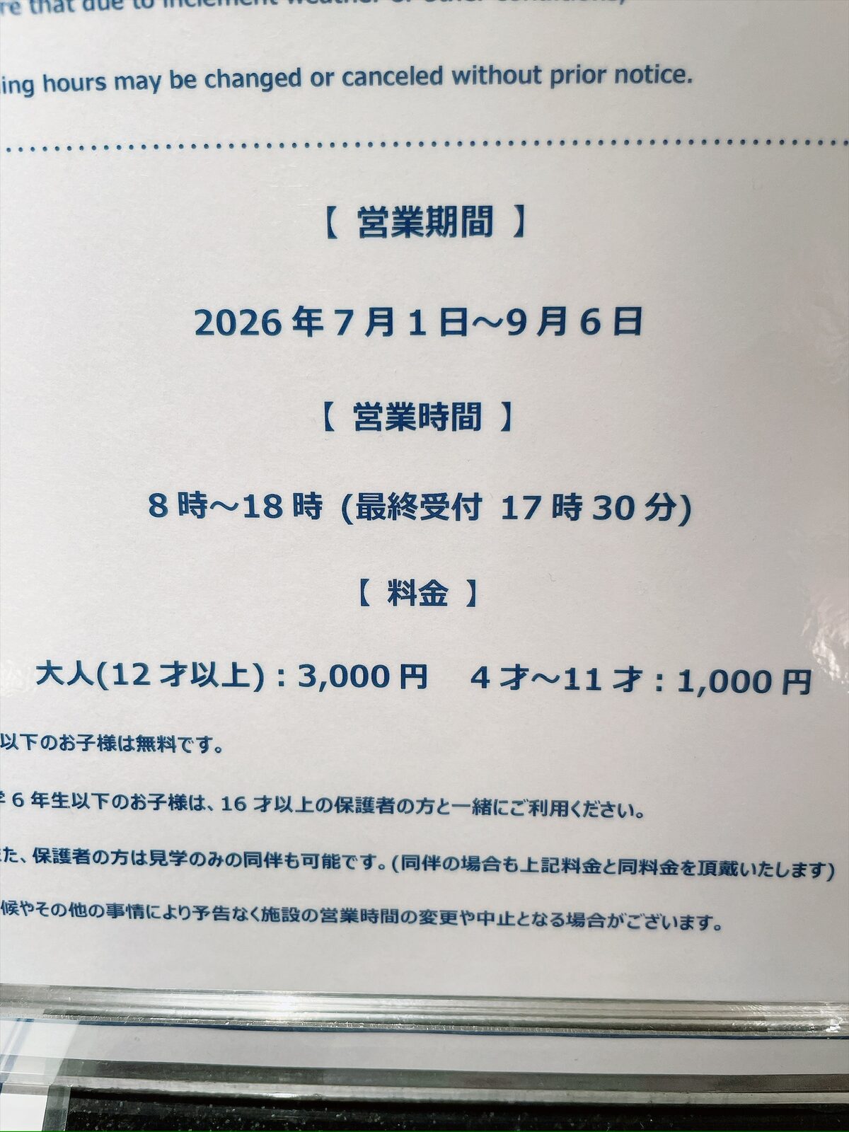プールの案内板のアップ。「営業期間 2026年7月1日〜9月6日」「営業時間 8時〜18時(最終受付17時30分)」「料金 大人(12才以上):3,000円 4才〜11才:1,000円」と書かれ、その下に同伴や変更についての注意書きが続く
