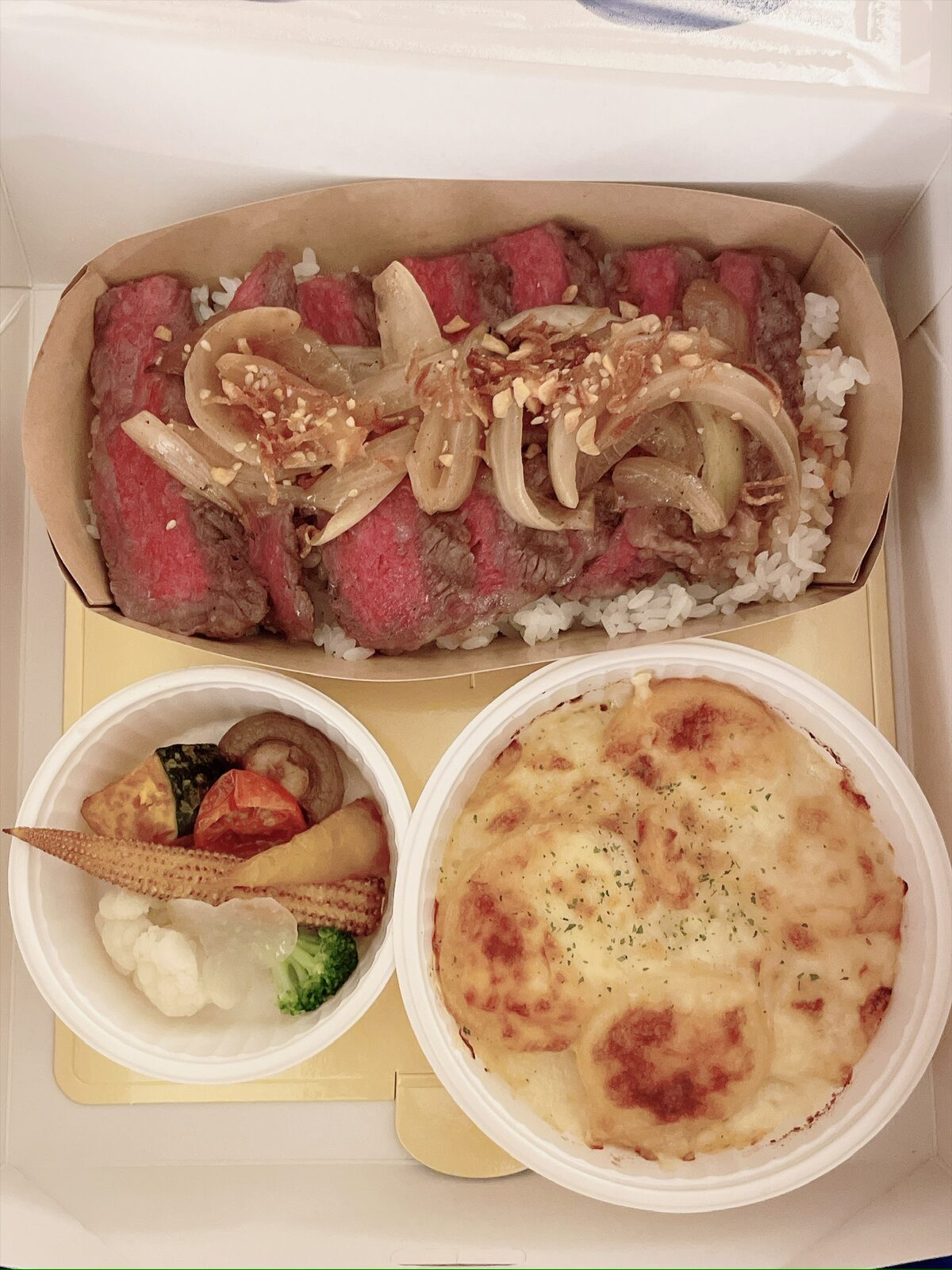 白い箱を開けたところ。紙の器に、ごはんの上へ薄切りのステーキを並べ、炒めた玉ねぎとくだいたナッツをのせた丼。手前に焼き色のついたポテトグラタンと、ヤングコーン・カリフラワー・ブロッコリー・かぼちゃなどのグリル野菜