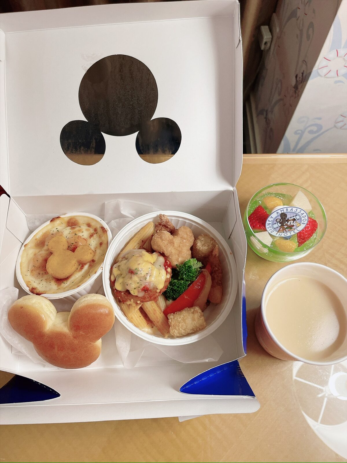 白い箱を開けたところ。ミッキーの形のパン、ミッキーの形のチップがのったグラタン、ペンネとハンバーグ・からあげ・ブロッコリーの入った器が並ぶ。箱のふたにはミッキーの形の穴。右に、いちごと角切りの果物が入った緑のゼリーと、紙コップのジュース