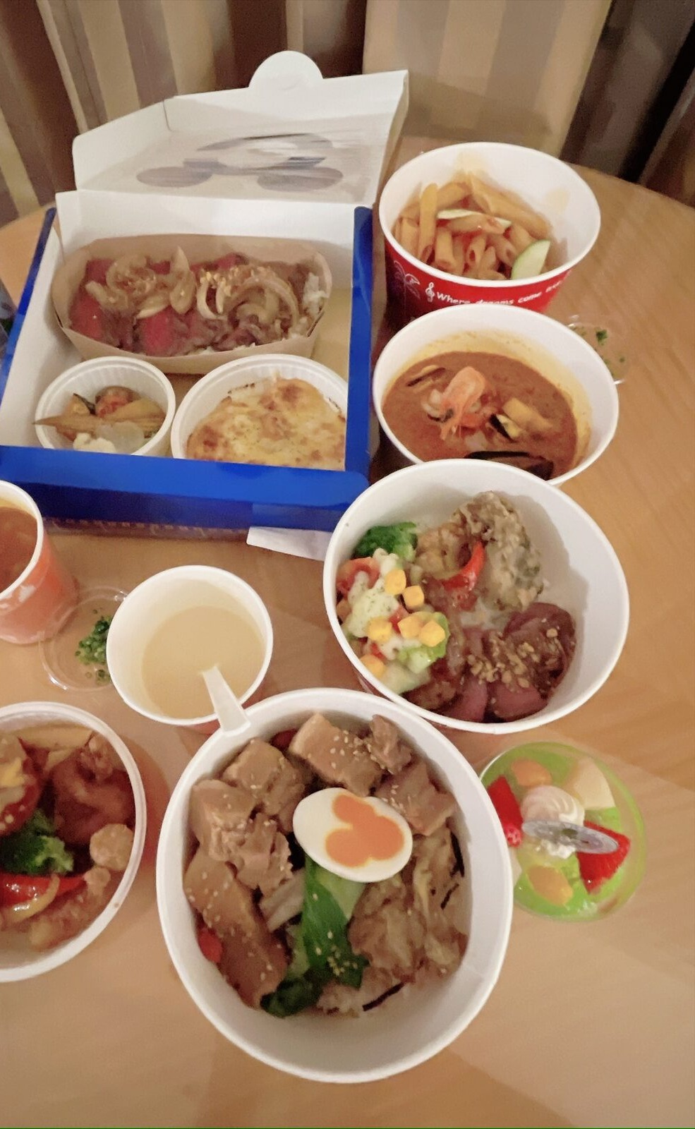 客室の木の丸テーブルに並んだ持ち帰りの料理。青いふちの箱の中にステーキ丼・グラタン・野菜、そのまわりに赤いカップのペンネ、えびの入った赤いスープ、白い丼が2つ、緑のゼリー、スープのカップが置かれている