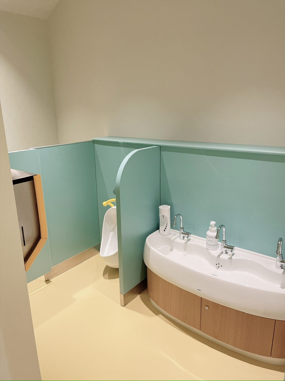 水色の壁の子ども用トイレ。低い位置に、蛇口が3つ並んだ横長の白い洗面台。左奥に小さな小便器と水色の仕切り、壁にペーパータオルの箱がある