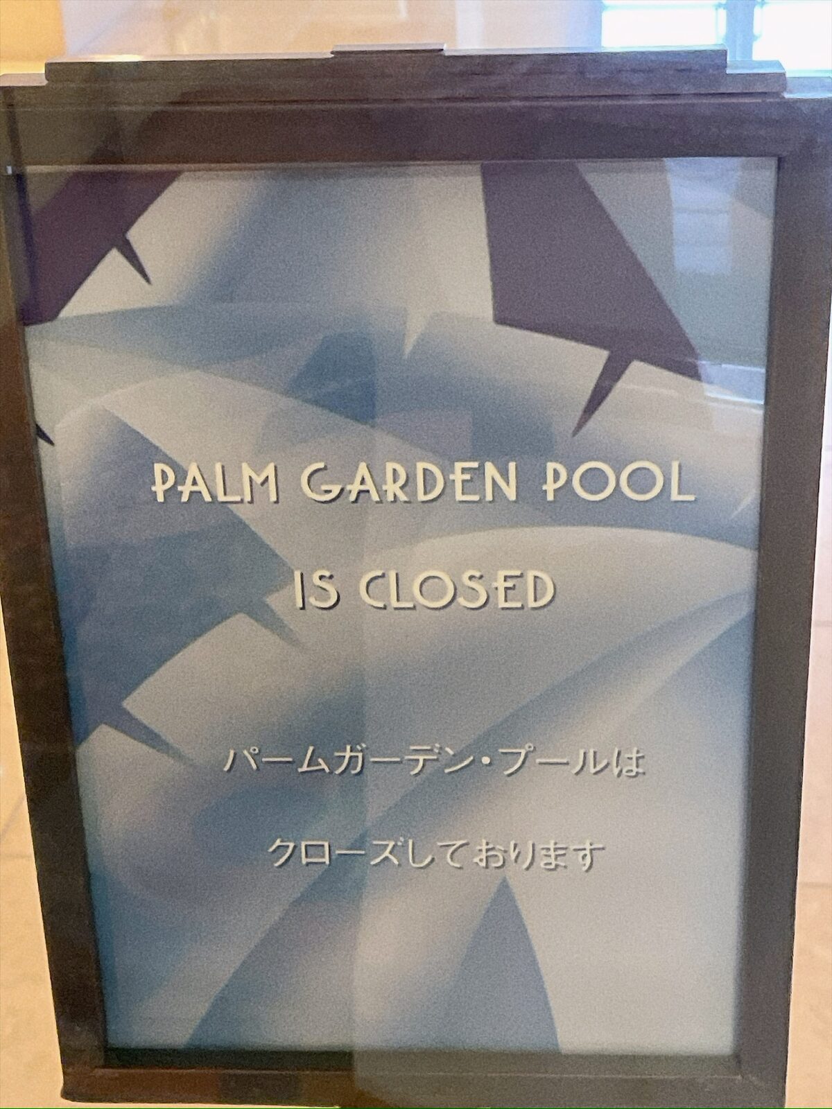 黒い枠のスタンドに入った掲示。水色の幾何学模様の地に「PALM GARDEN POOL IS CLOSED」「パームガーデン・プールは クローズしております」と白い文字で書かれている