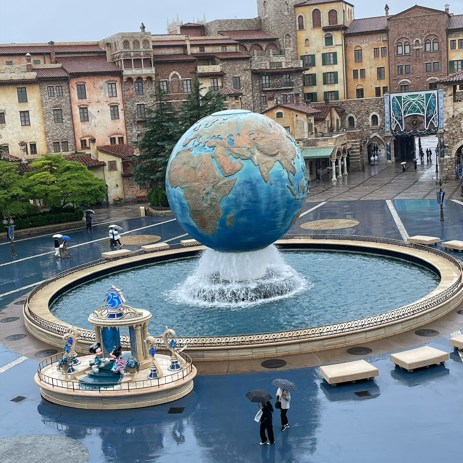 高いところから見下ろしたディズニーシーの入口。手前に25周年のモニュメント、その奥に地球儀と噴水。地面が濡れ、傘をさした人が歩いている
