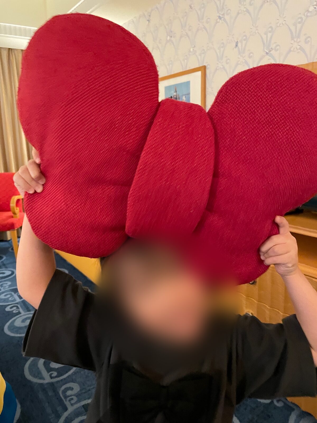 赤い大きな蝶ネクタイの形のクッションを、子どもが両手で頭の上に持ち上げている。奥は客室の壁紙と額に入った絵(子どもの顔と髪はぼかし処理)