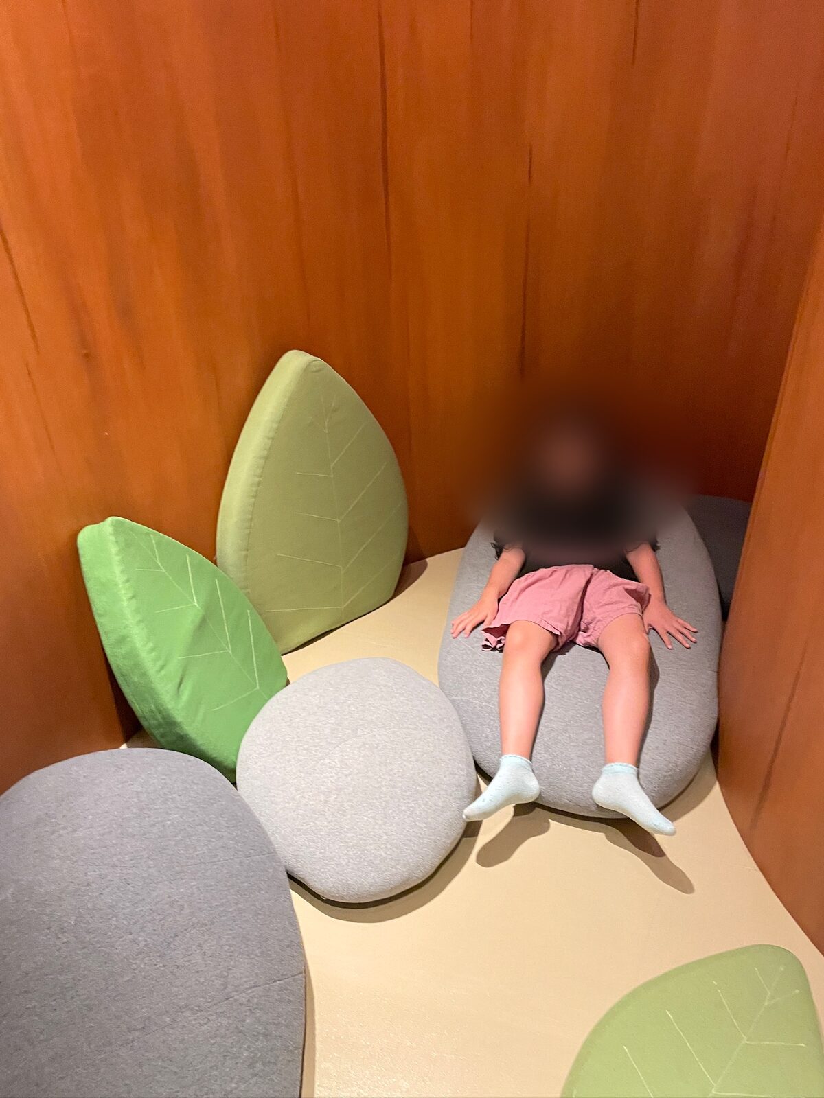 木の幹の内側のような、オレンジ色の板に囲まれたくぼみ。灰色の石の形のクッションと、緑の葉の形のクッションが並び、子どもが仰向けに寝そべっている(子どもの顔と髪はぼかし処理)