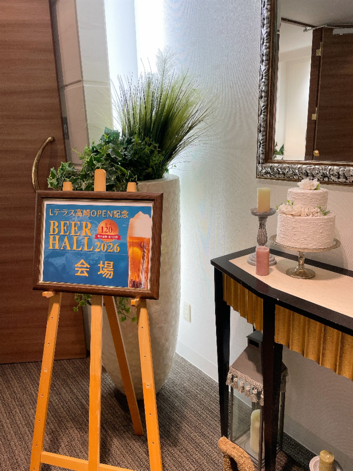 イーゼルに立てられた「Lテラス高崎OPEN記念 BEER HALL 2026 会場」の看板。横のテーブルにウェディングケーキの飾り