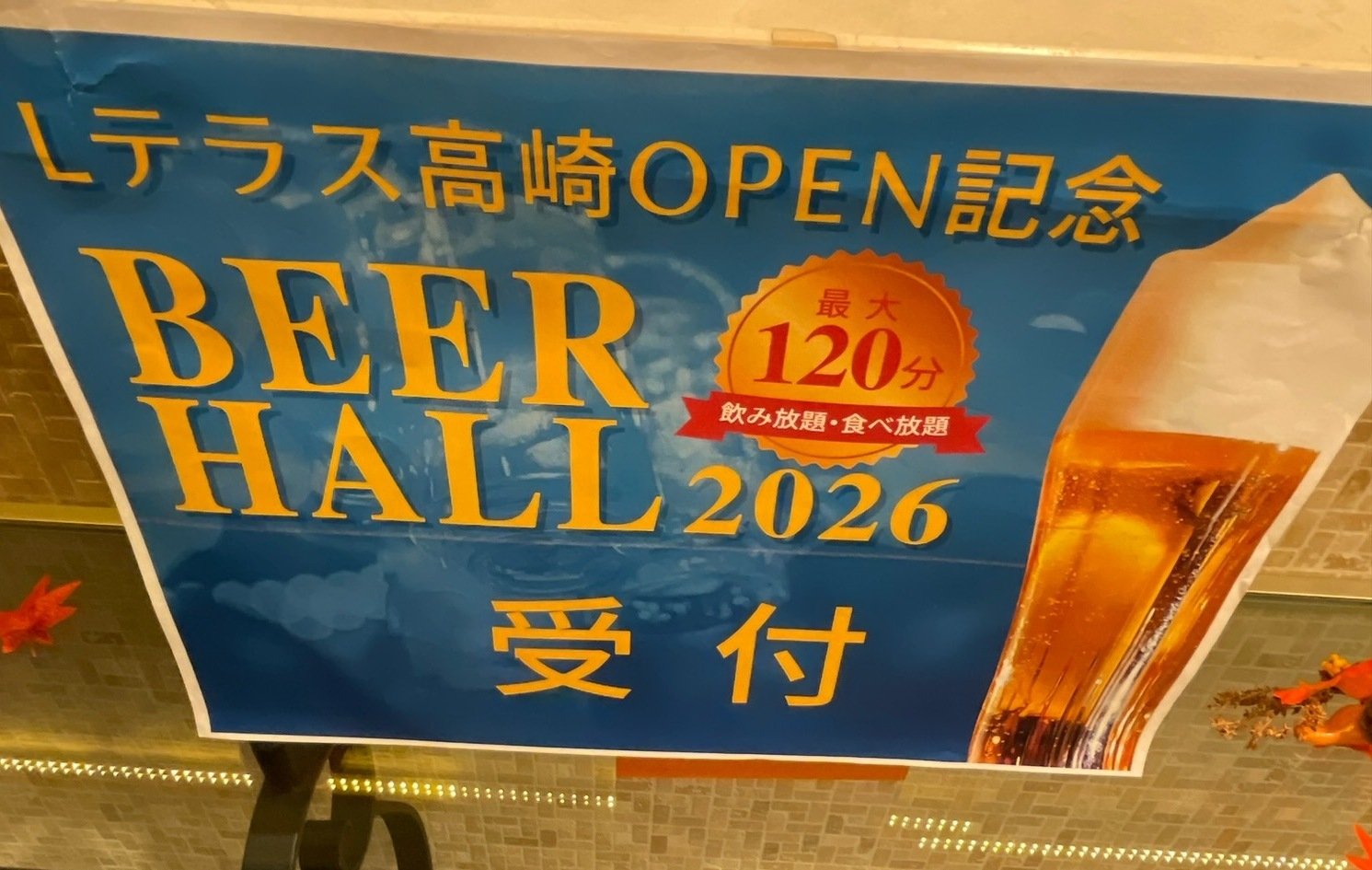 「Lテラス高崎OPEN記念 BEER HALL 2026 受付」と書かれた青いポスター。ビールのジョッキの写真と「最大120分 飲み放題・食べ放題」の文字