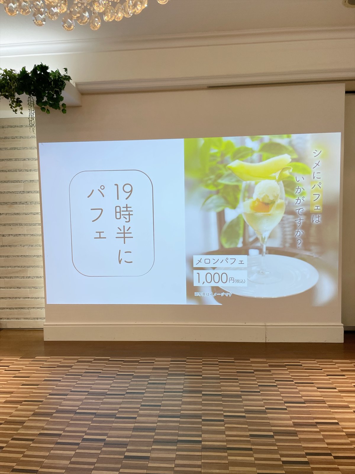 会場のスクリーンに映された案内。「19時半にパフェ」「シメにパフェはいかがですか？」「メロンパフェ 1,000円（税込）」の文字