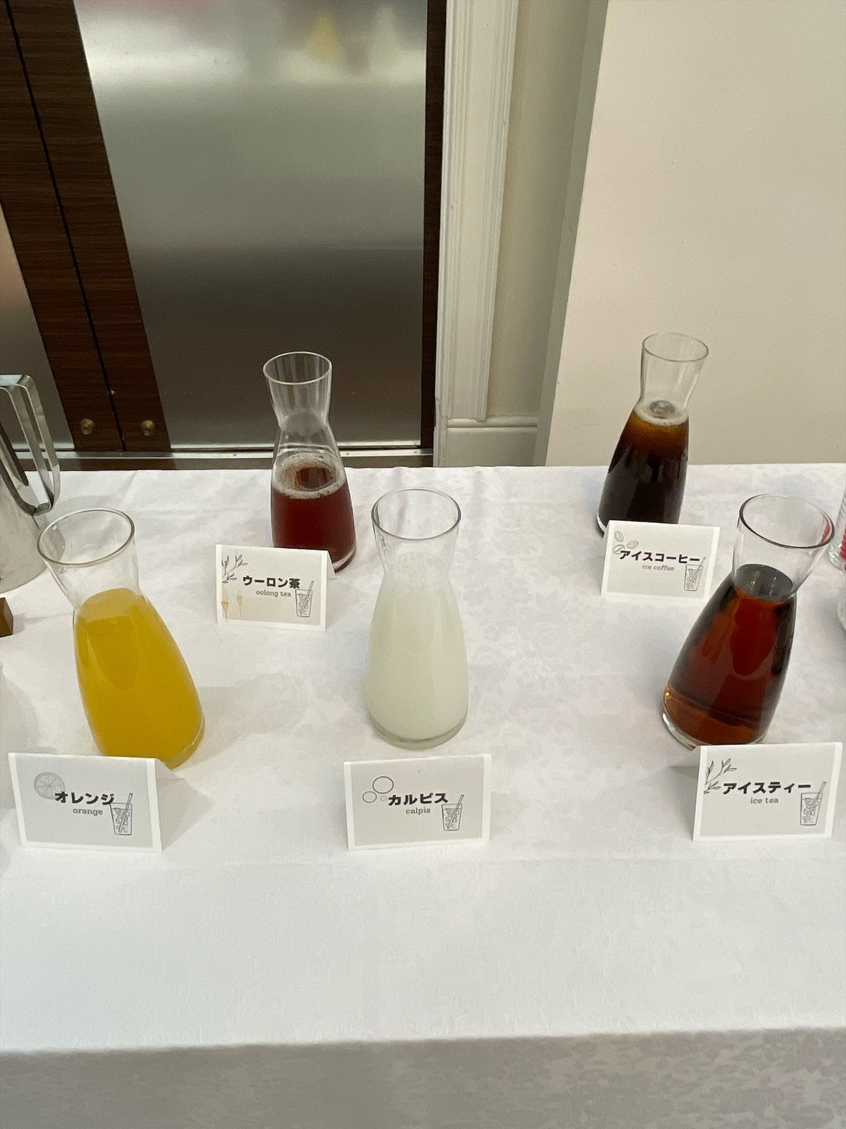 白いクロスの台に、ガラスの水差しが5本。オレンジ、ウーロン茶、カルピス、アイスコーヒー、アイスティーの名札が立っている