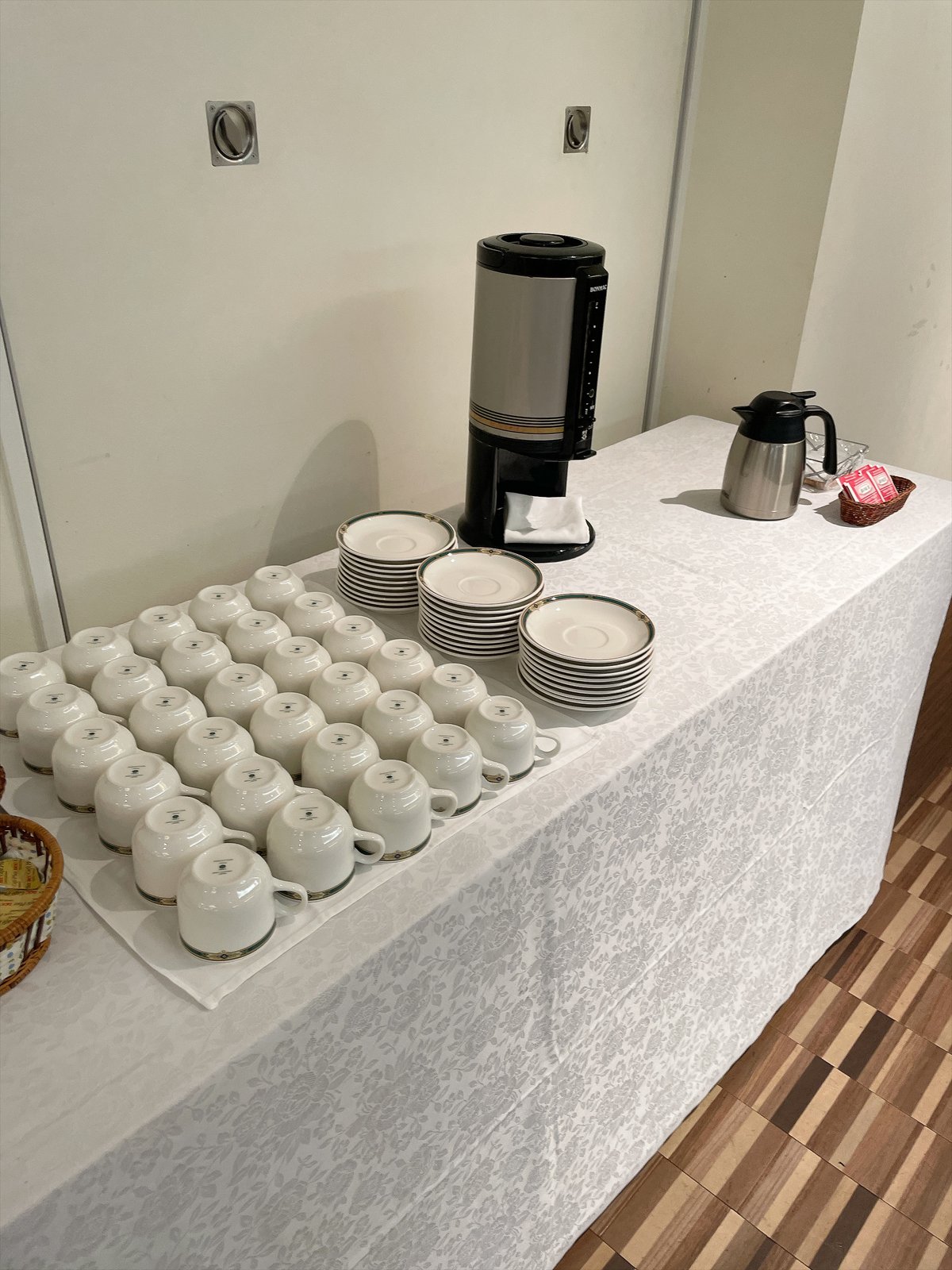 白いクロスの台に、白いコーヒーカップとソーサーが並び、奥にコーヒーのポット