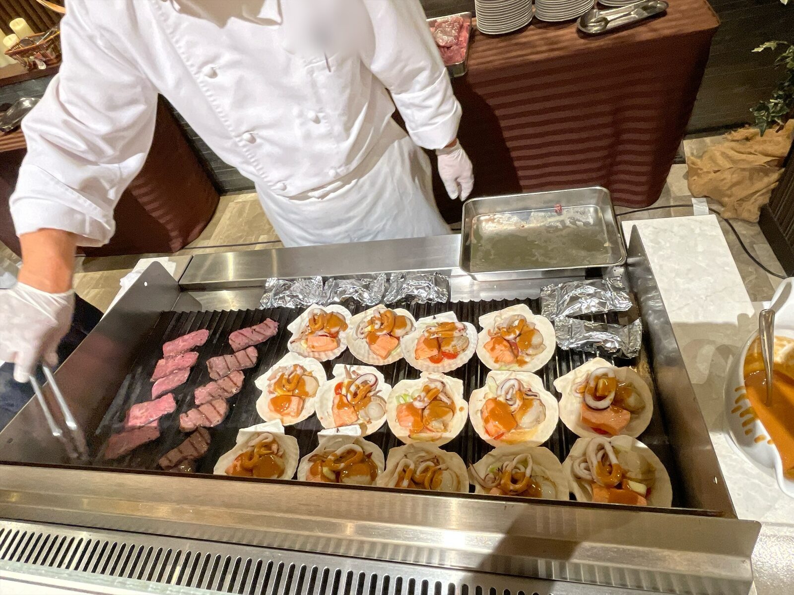 コックコート姿のシェフが、鉄板でステーキとホタテの貝殻に盛った魚介を焼いている。奥にアルミホイルの包みが並ぶ