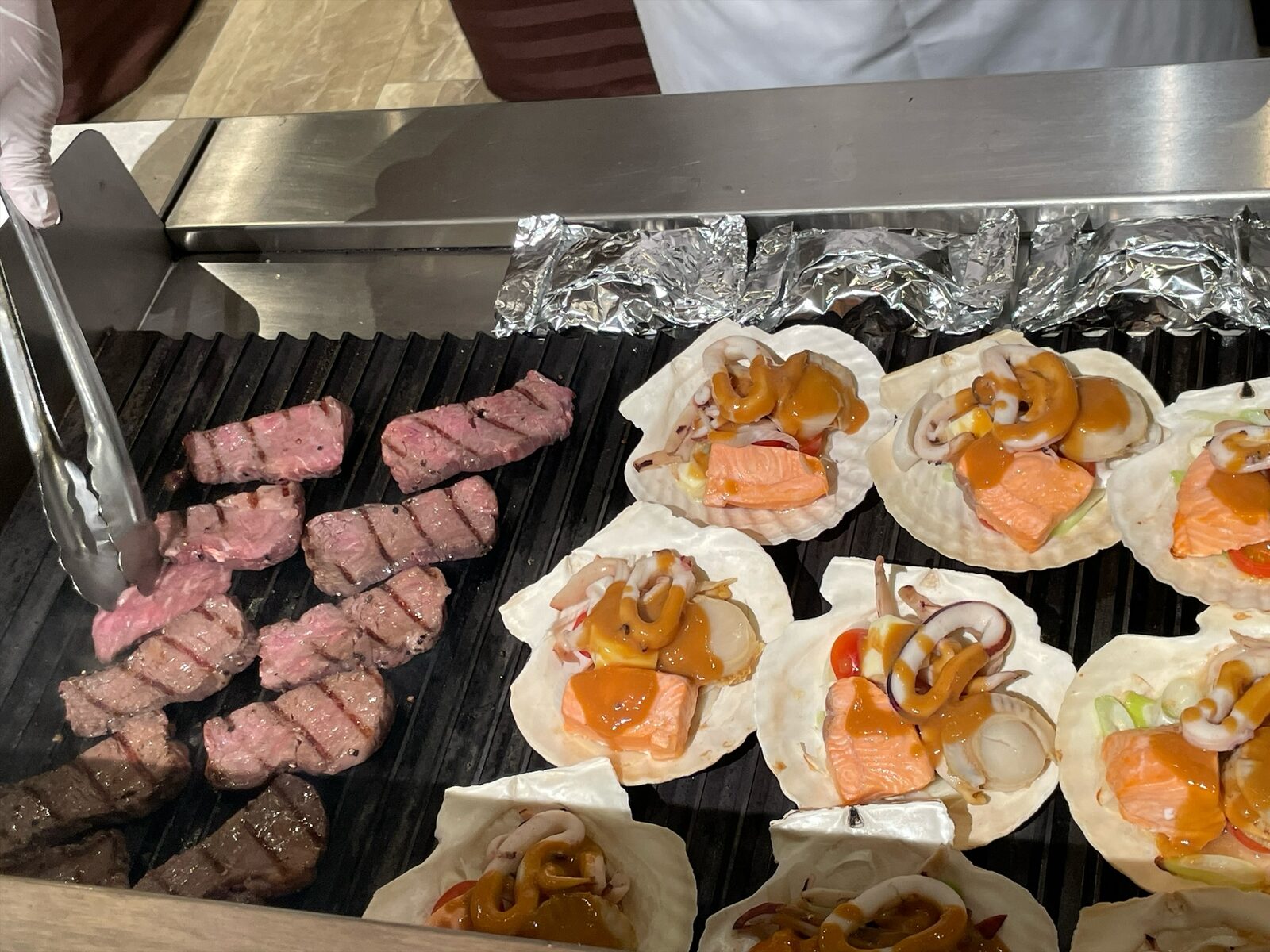 鉄板の左で焼かれる牛肉のステーキと、右に並ぶホタテの貝殻に盛られた魚介の焼き物。奥にアルミホイルの包み