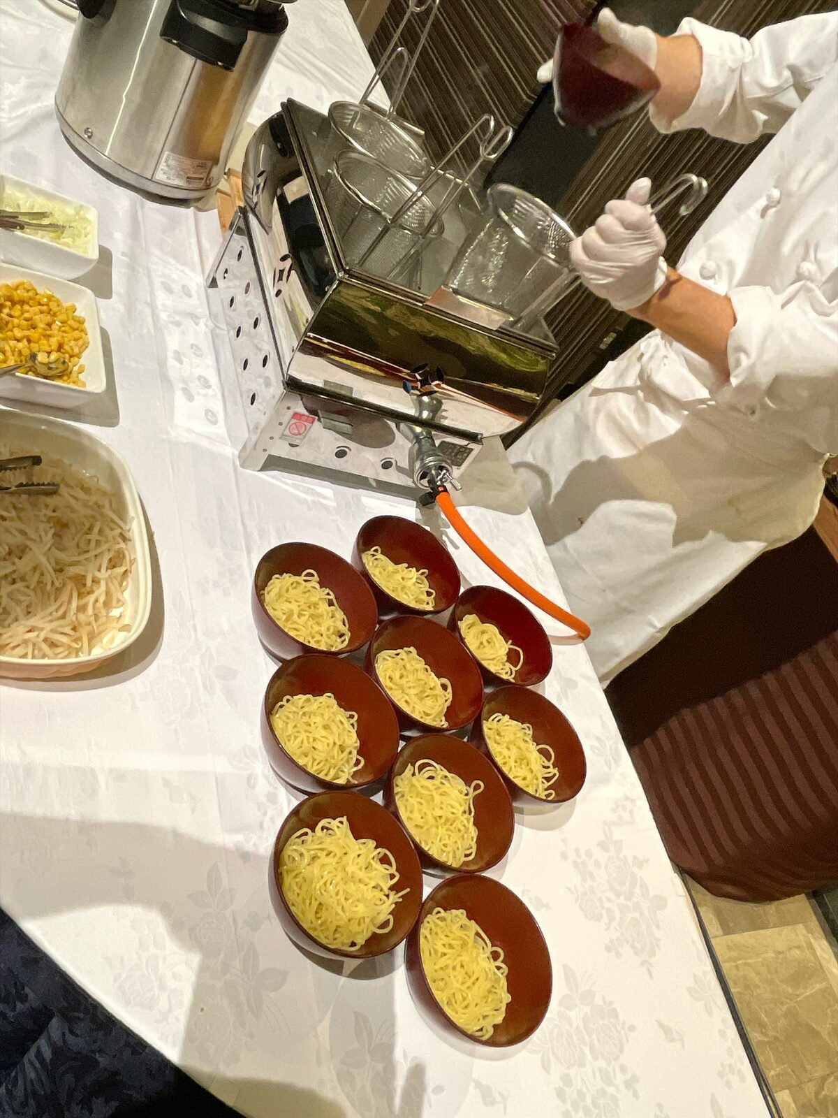 白いクロスの台に、赤い漆のお椀9つに1人分ずつ分けられたラーメンの麺。奥にコーンともやし