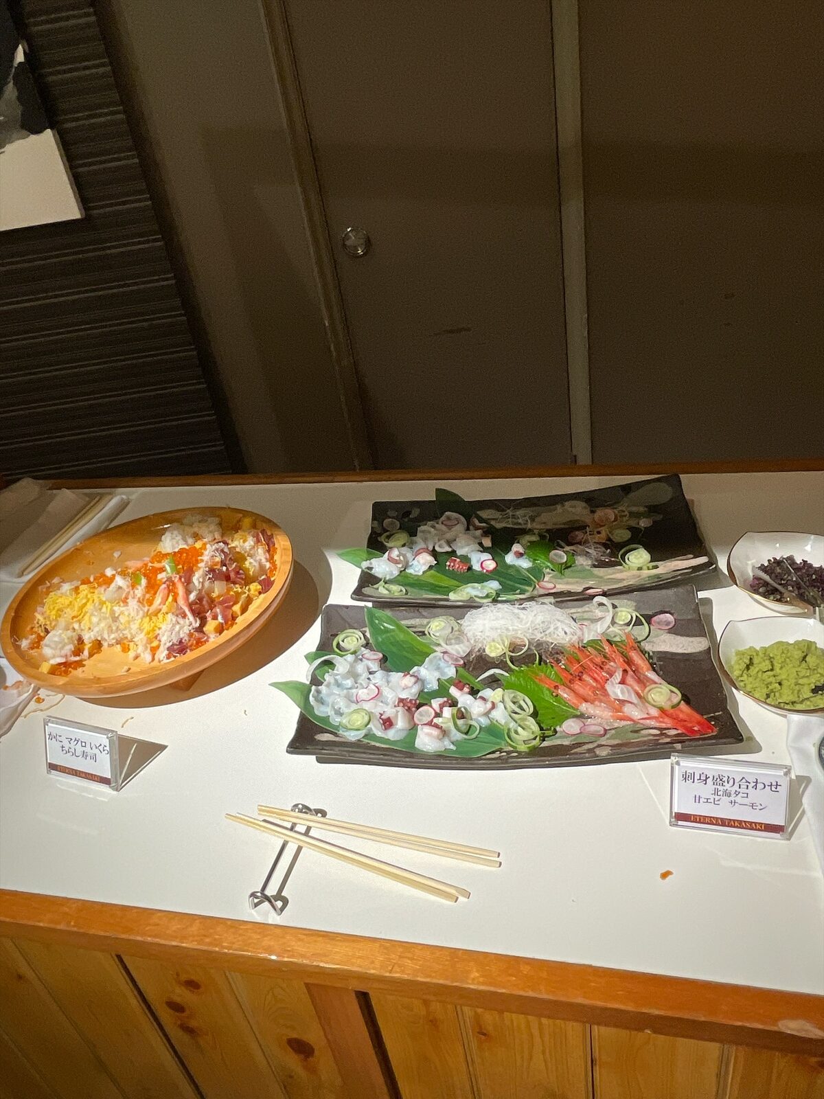 木のカウンターに、黒い角皿の刺身盛り合わせと、木の器のちらし寿司。食札とわさびが添えられている