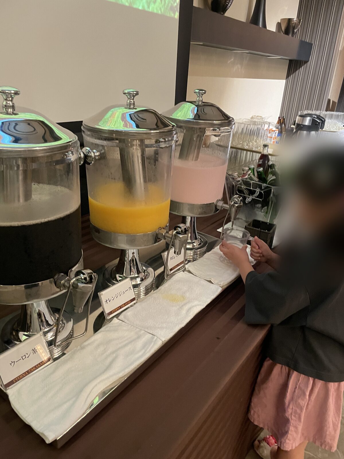 子どもが自分でカップを持ち、ピンク色の飲みもののディスペンサーから注いでいる
