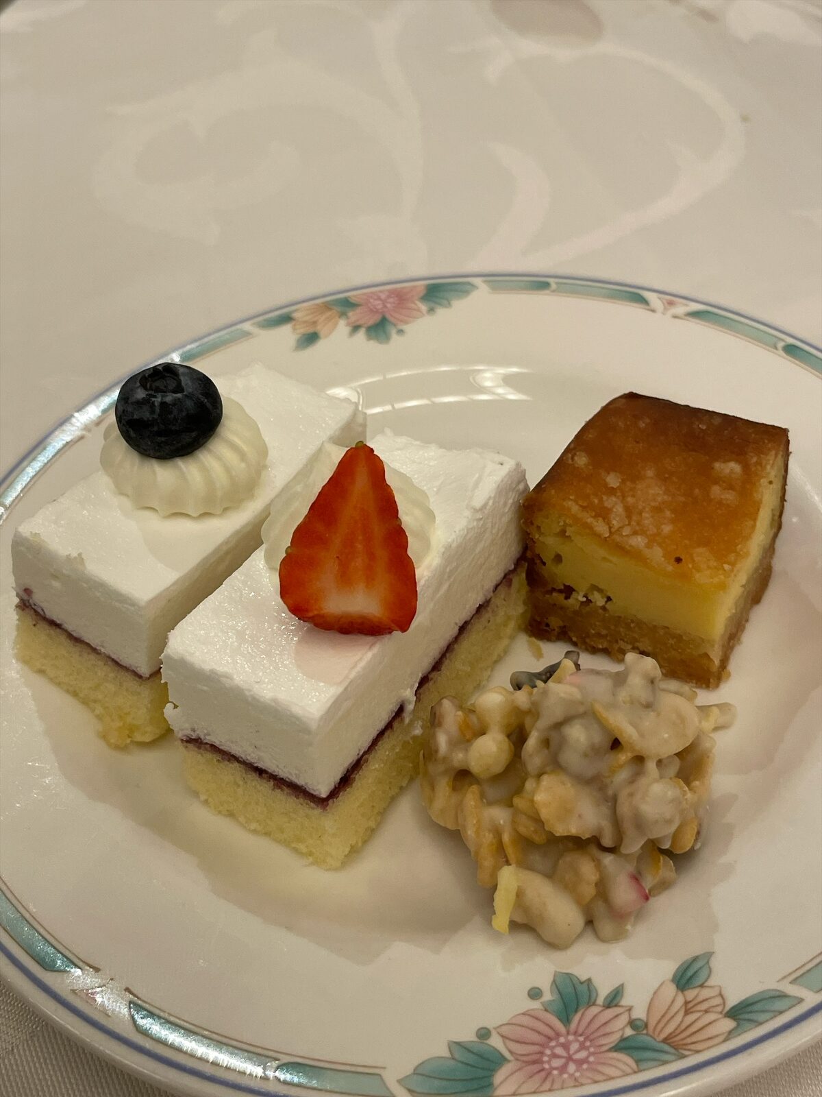 花柄の皿に、レアチーズケーキ2切れ、焼き色のついたチーズの焼き菓子、白いチョコでからめたシリアル菓子