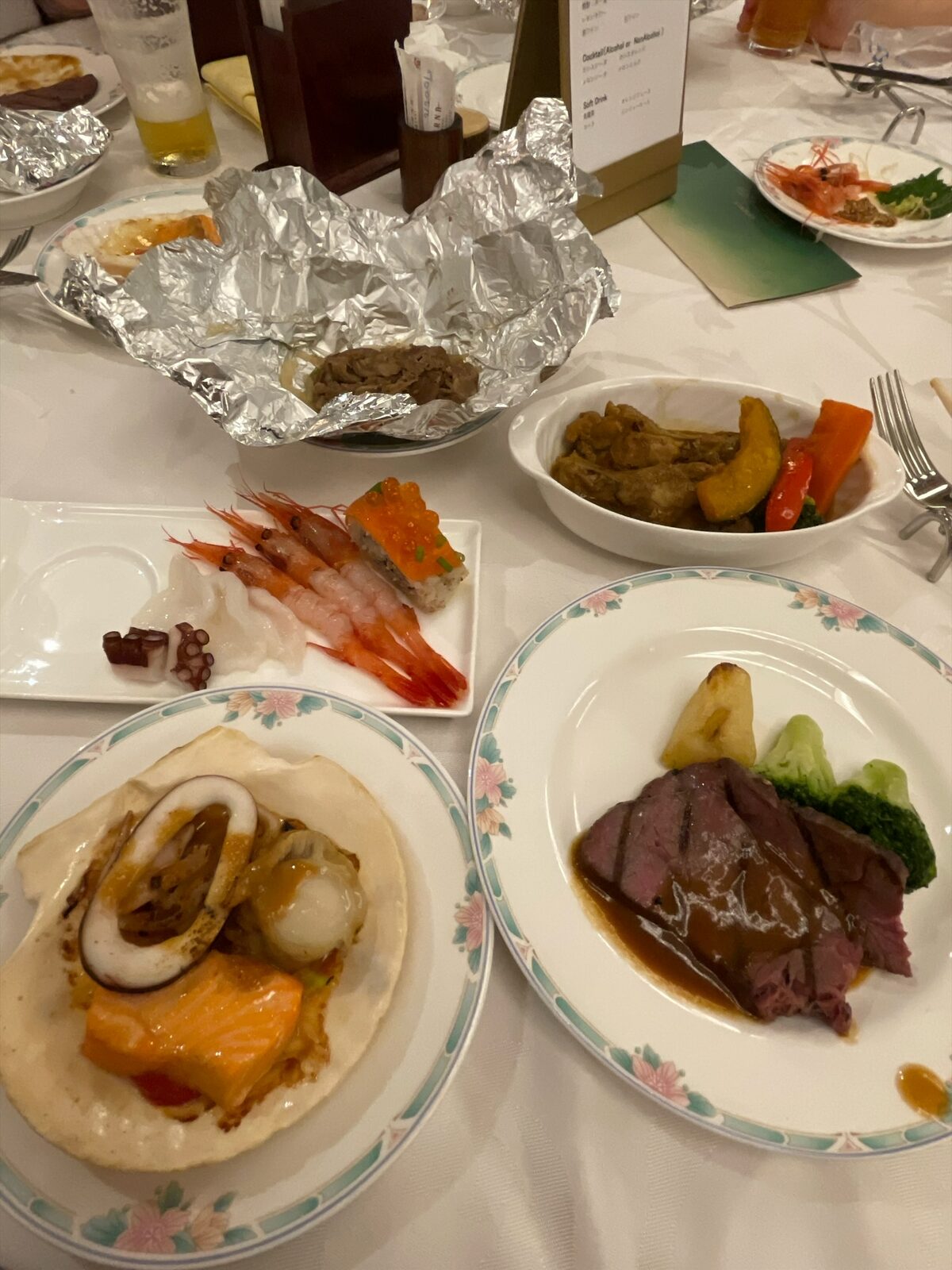 テーブルに並んだ取り皿。ホタテの貝焼き、ソースのかかったステーキ、刺身、スープカレー、ホイルに包まれたジンギスカン