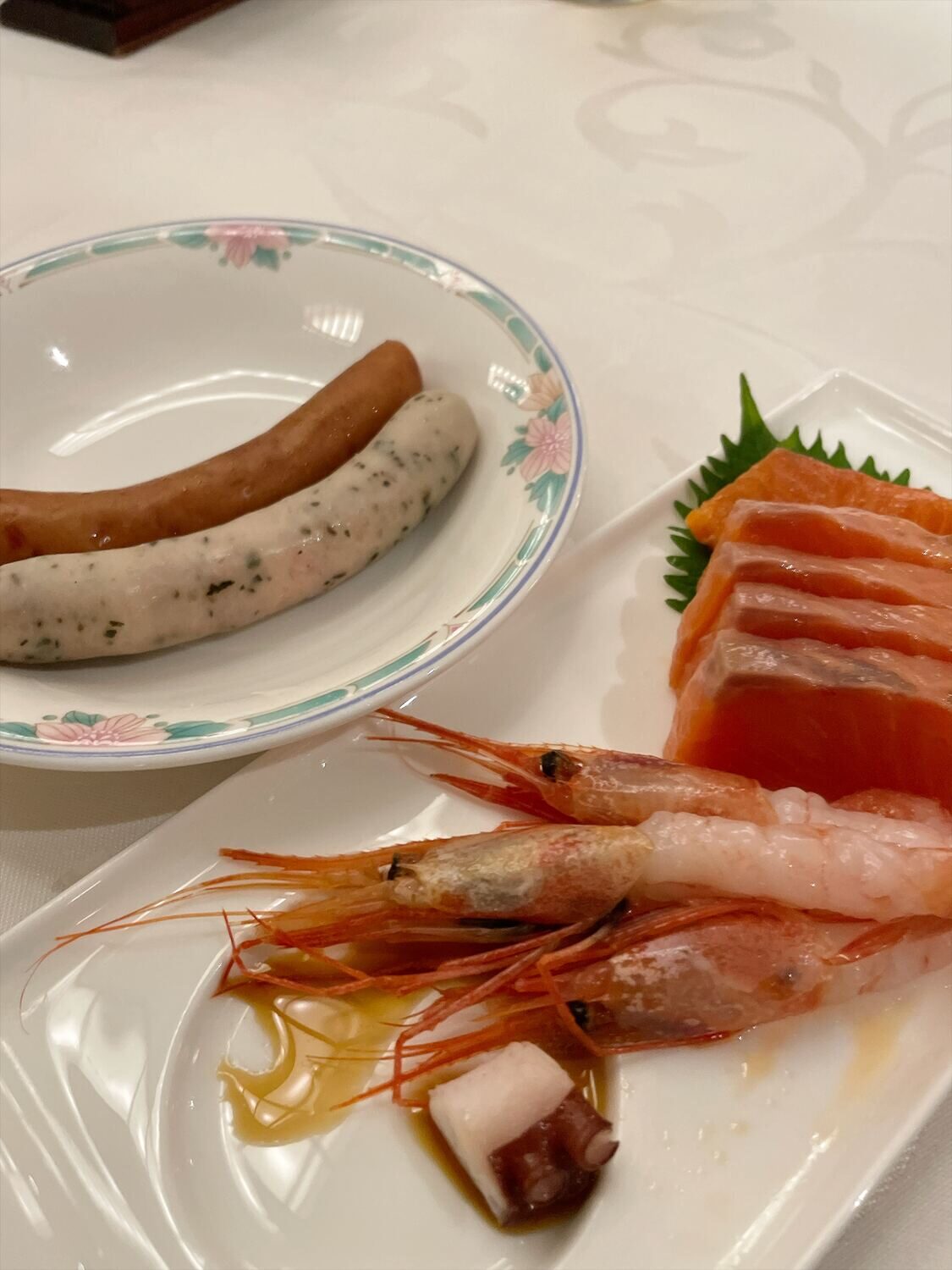 皿にのった2本のソーセージと、別皿のサーモンの刺身、頭つきの甘エビ、タコ